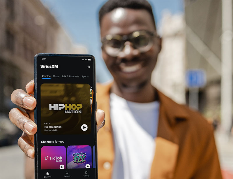 Man holding phone showing SiriusXM App with Hip Hop Nation, Tik Tok Radio and SiriusXM FLY